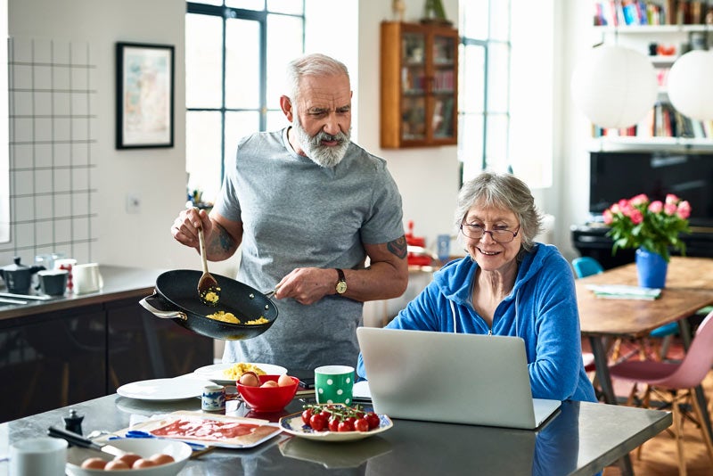 Senior woman using laptop and smiling as man serves breakfast